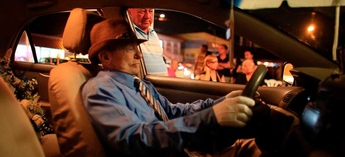 Een in zijn auto opgebaarde man zittend achter het stuur met een lichtblauw pak aan en een hoed op.